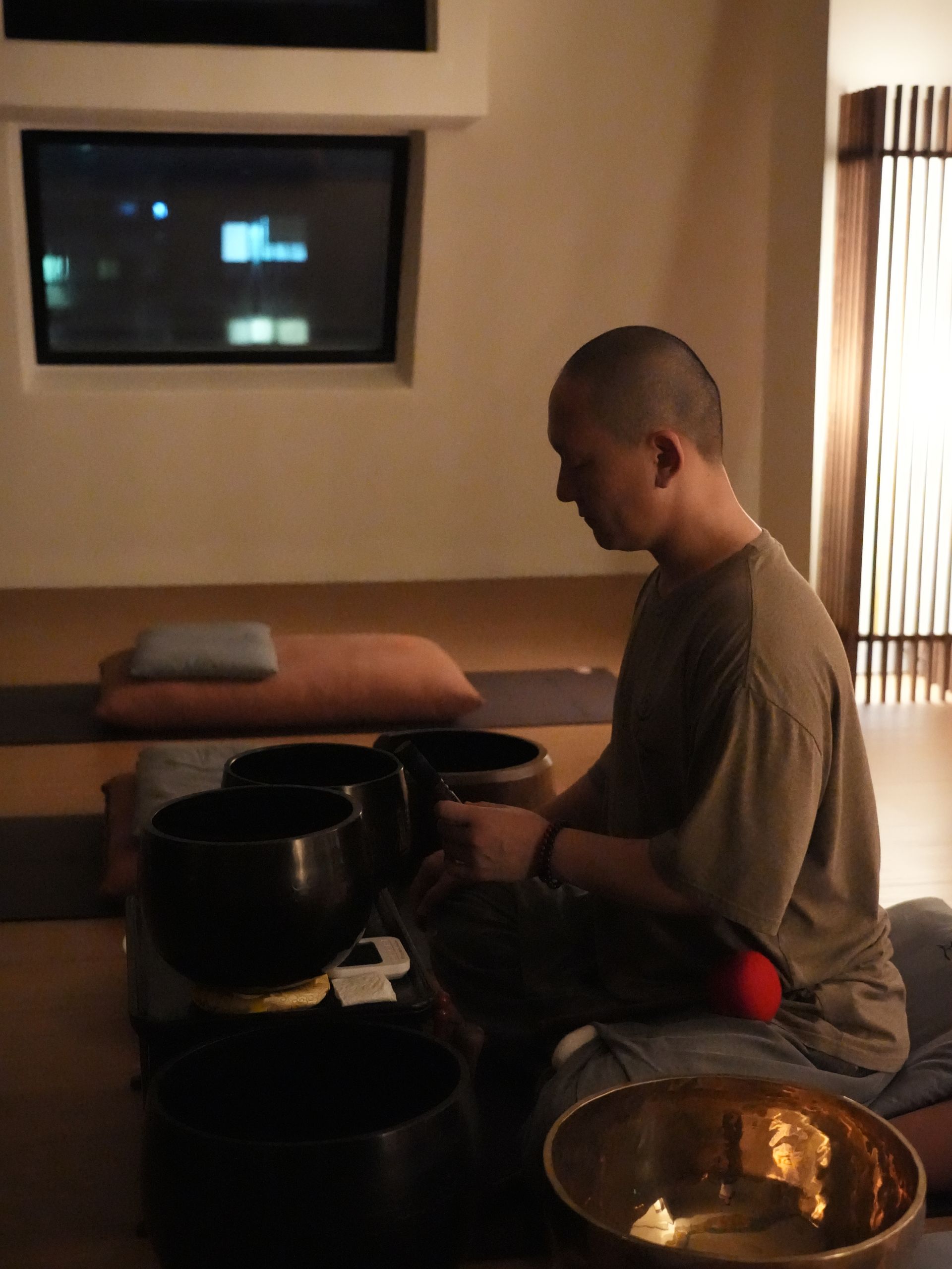 머리를 깎은 남자가 다리를 꼬고 앉아, 어두운 방에서 노래 그릇을 두드리고 있다.