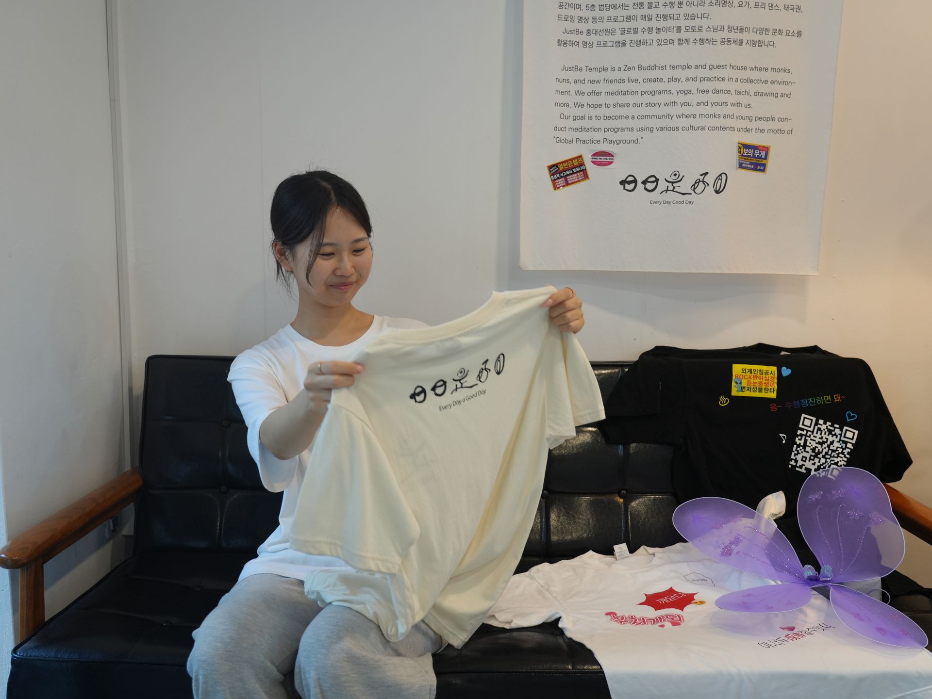 Woman holding up a cream-colored t-shirt, smiling. Other shirts and decorations on a couch against a white wall.
