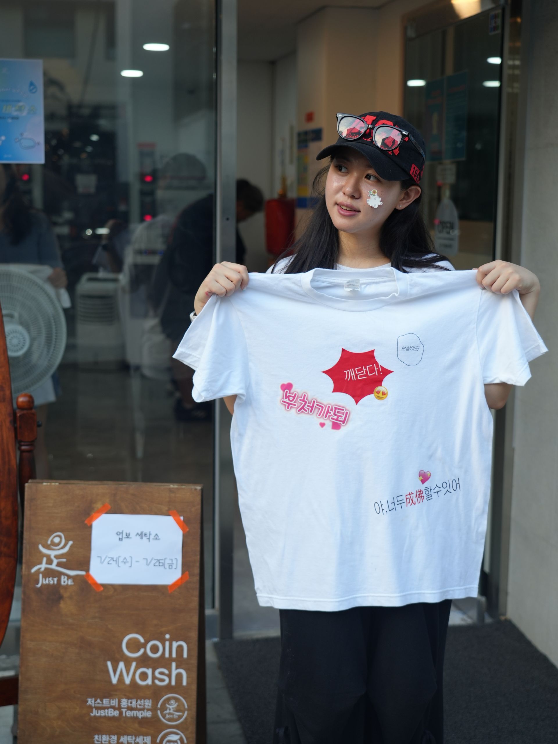 Woman holding up a white t-shirt with a red and pink design, standing in front of a laundromat.