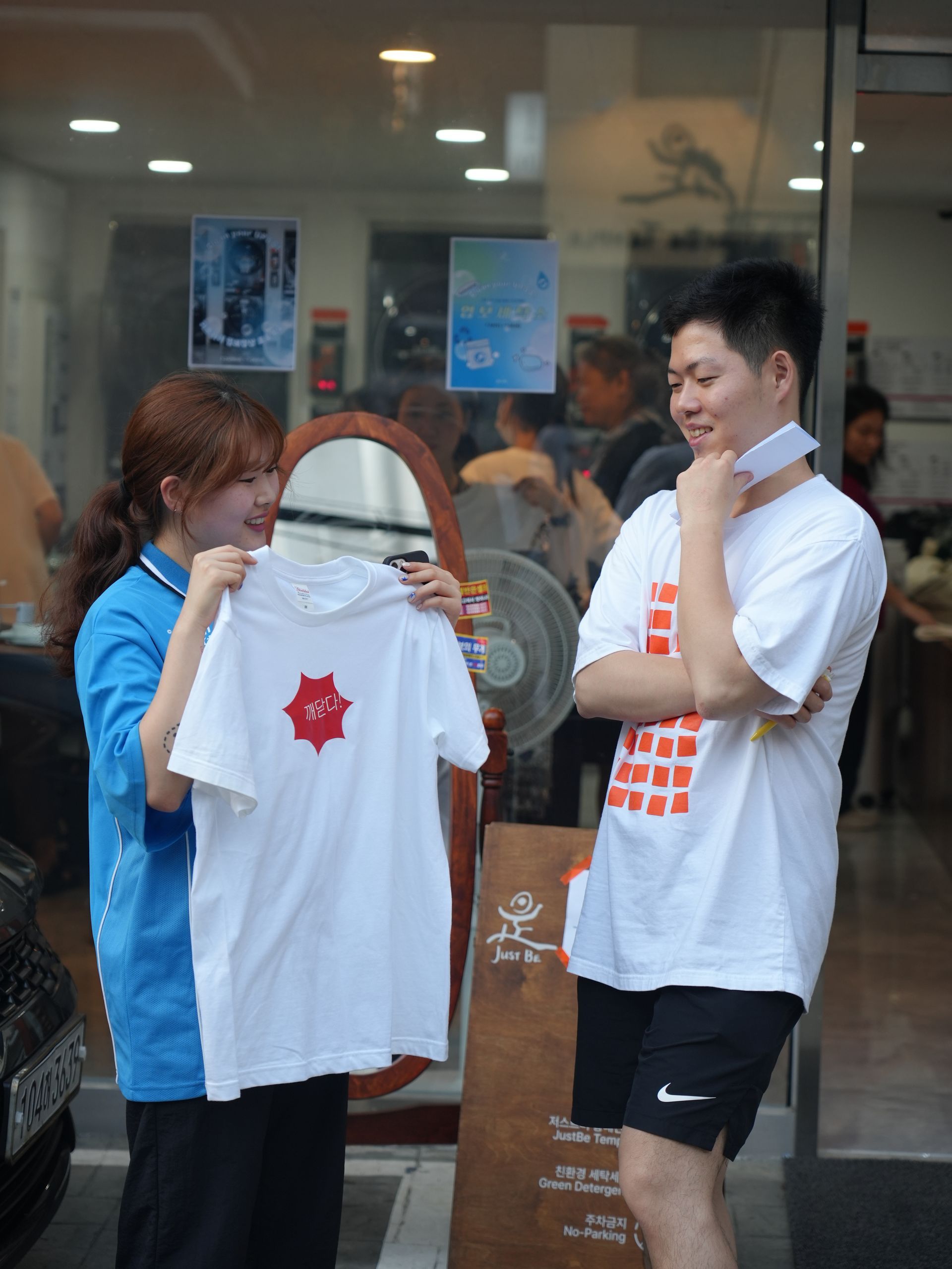 Woman showing a man a white t-shirt with a red design; outside a shop, both are smiling.