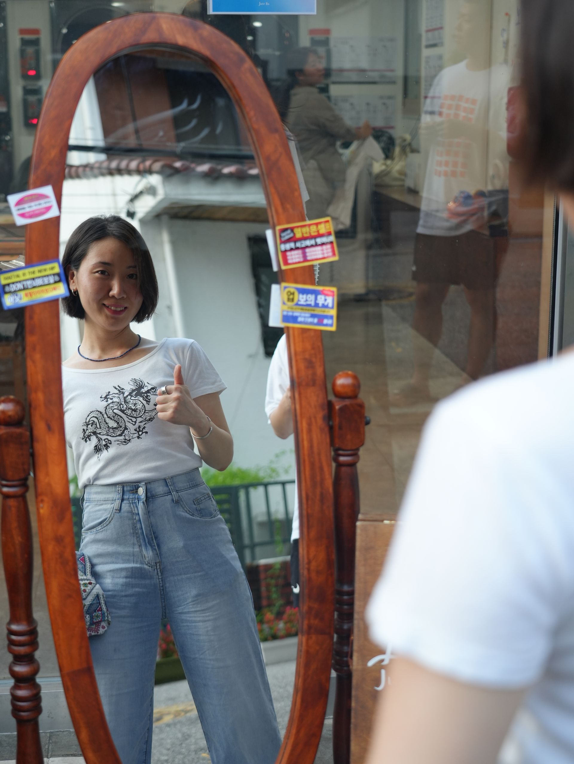 Woman in jeans and t-shirt giving thumbs up, reflected in a tall, wooden-framed mirror.