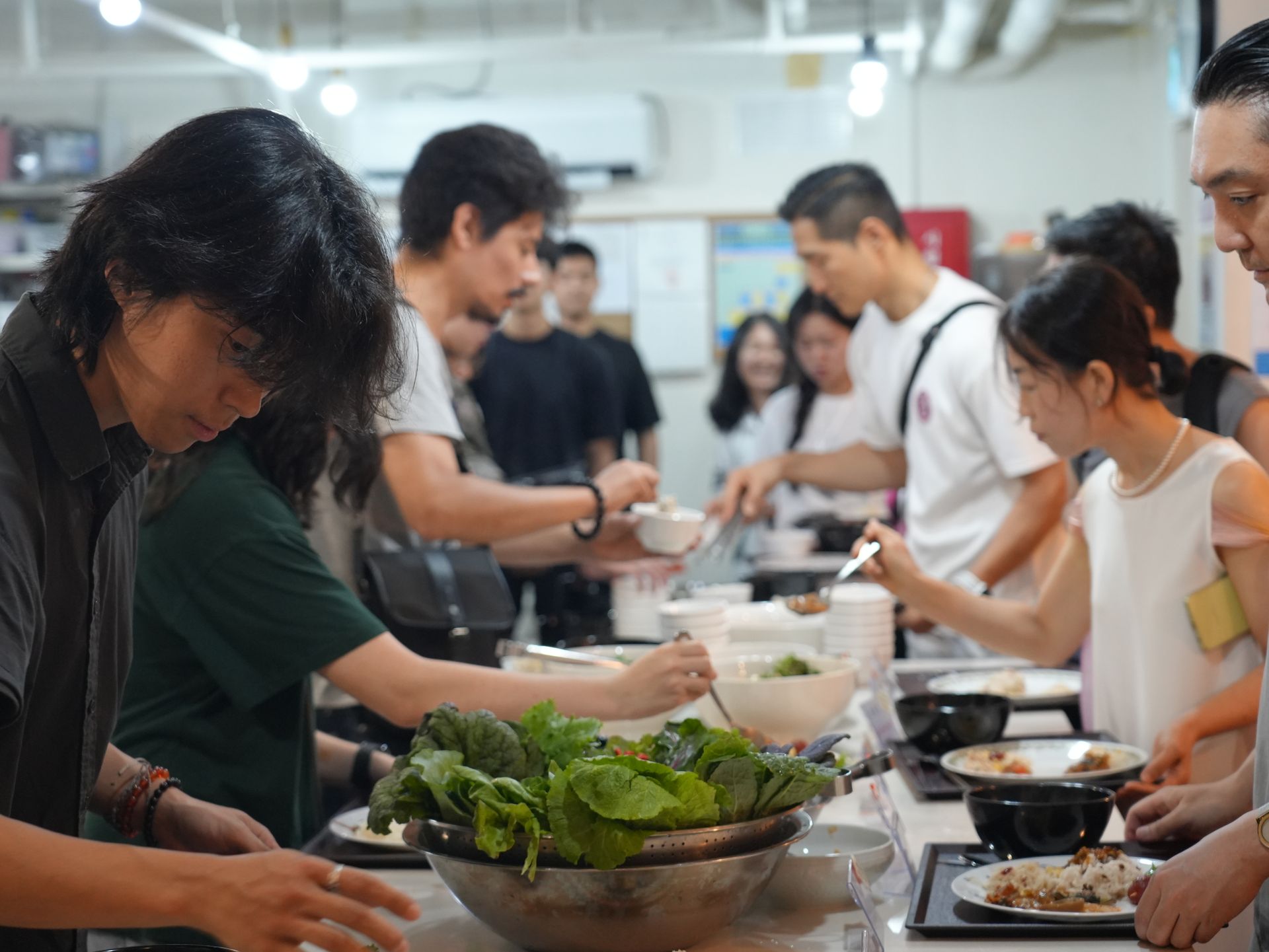 People serve themselves food at a buffet line. Many are reaching for bowls and greens in a brightly lit room.