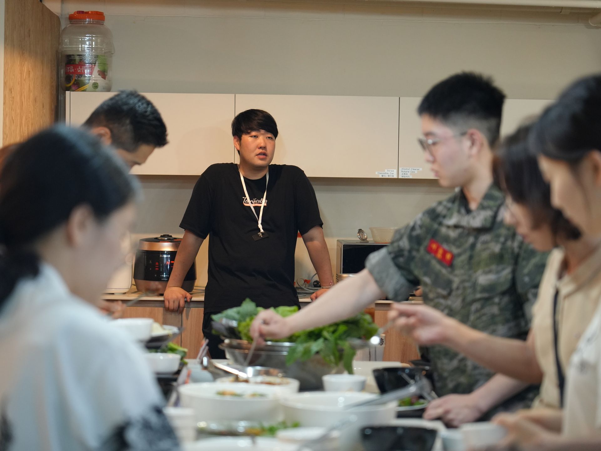 주방에서 음식을 준비하는 사람들, 검은 셔츠를 입은 남자 한 명, 군복을 입은 남자 한 명.