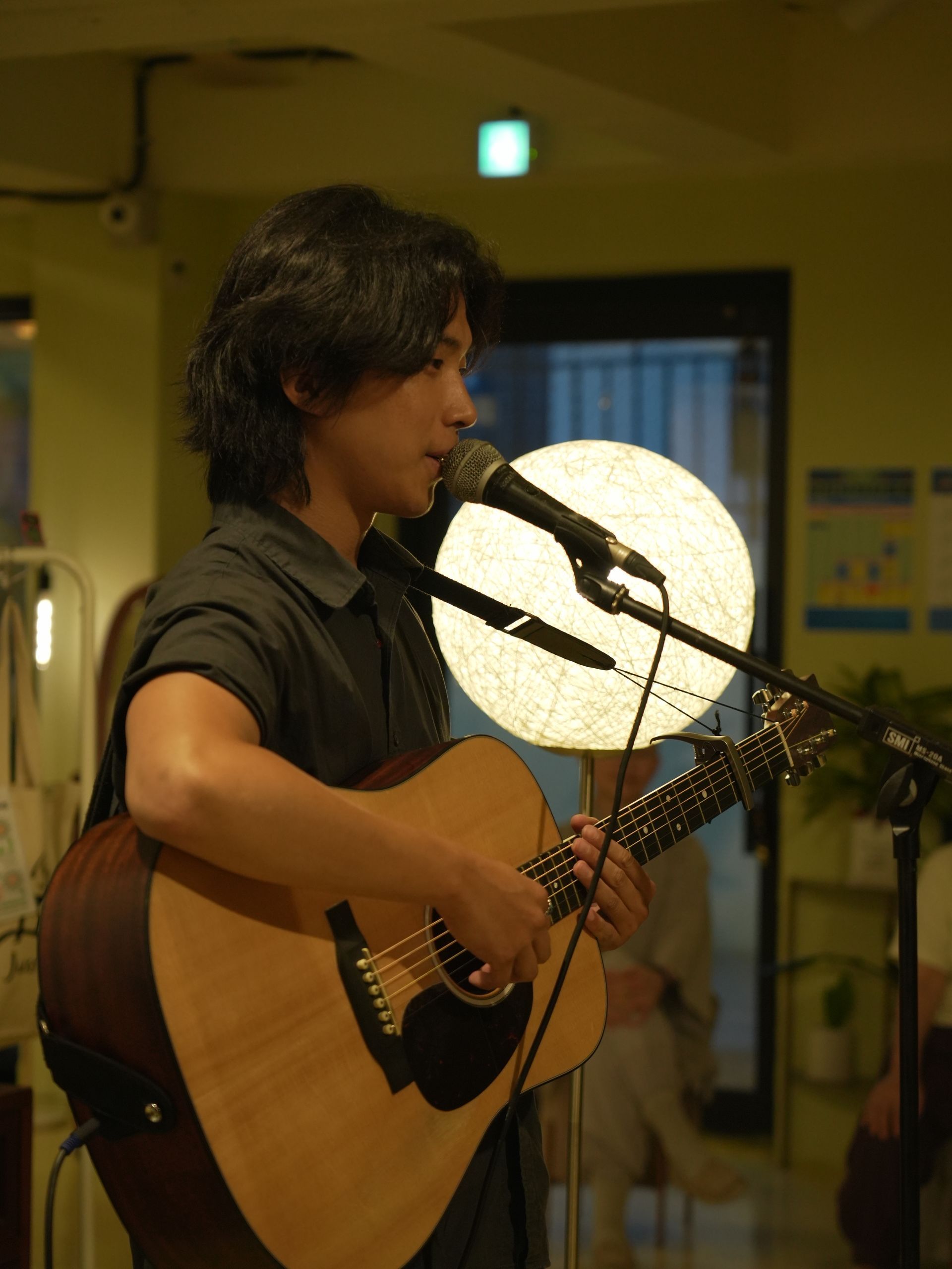 Man with long dark hair playing acoustic guitar, singing into a microphone, indoors.