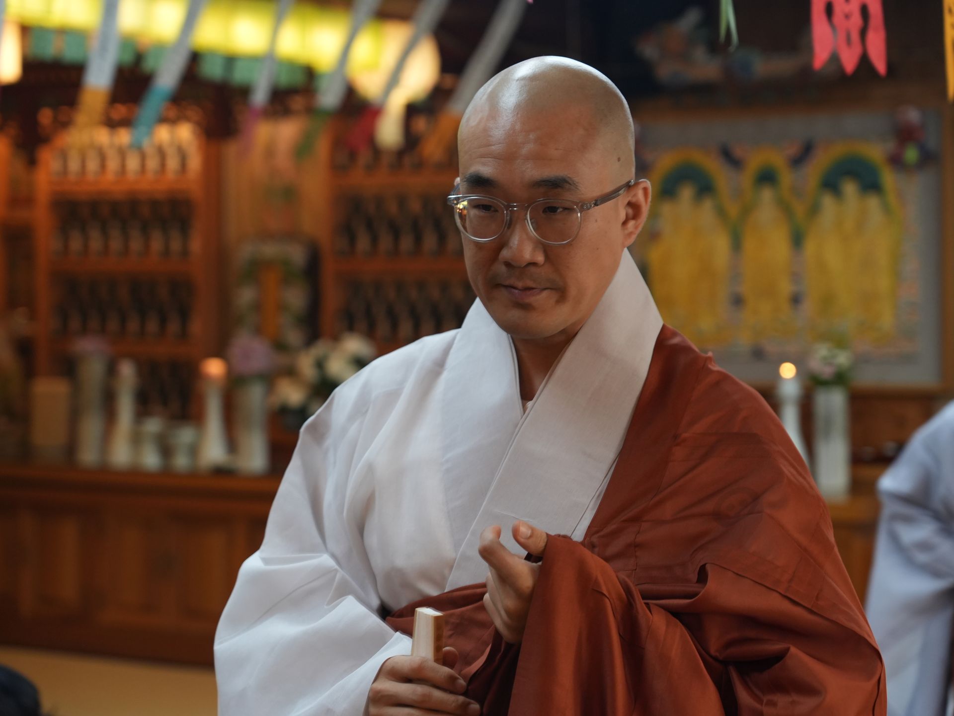 Bald monk in white and brown robes with glasses, in temple, holding a stick.