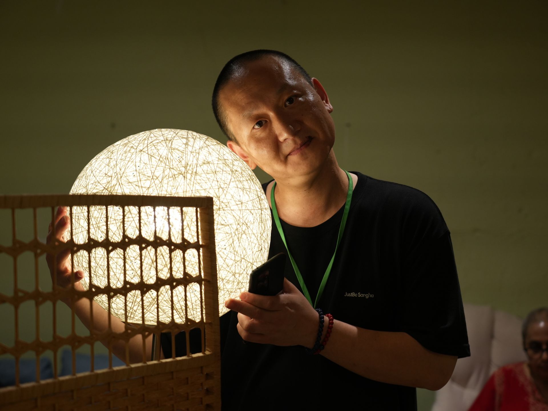 Man holding a lit, round, speckled lamp, with a rectangular wooden piece, indoors; he smiles.