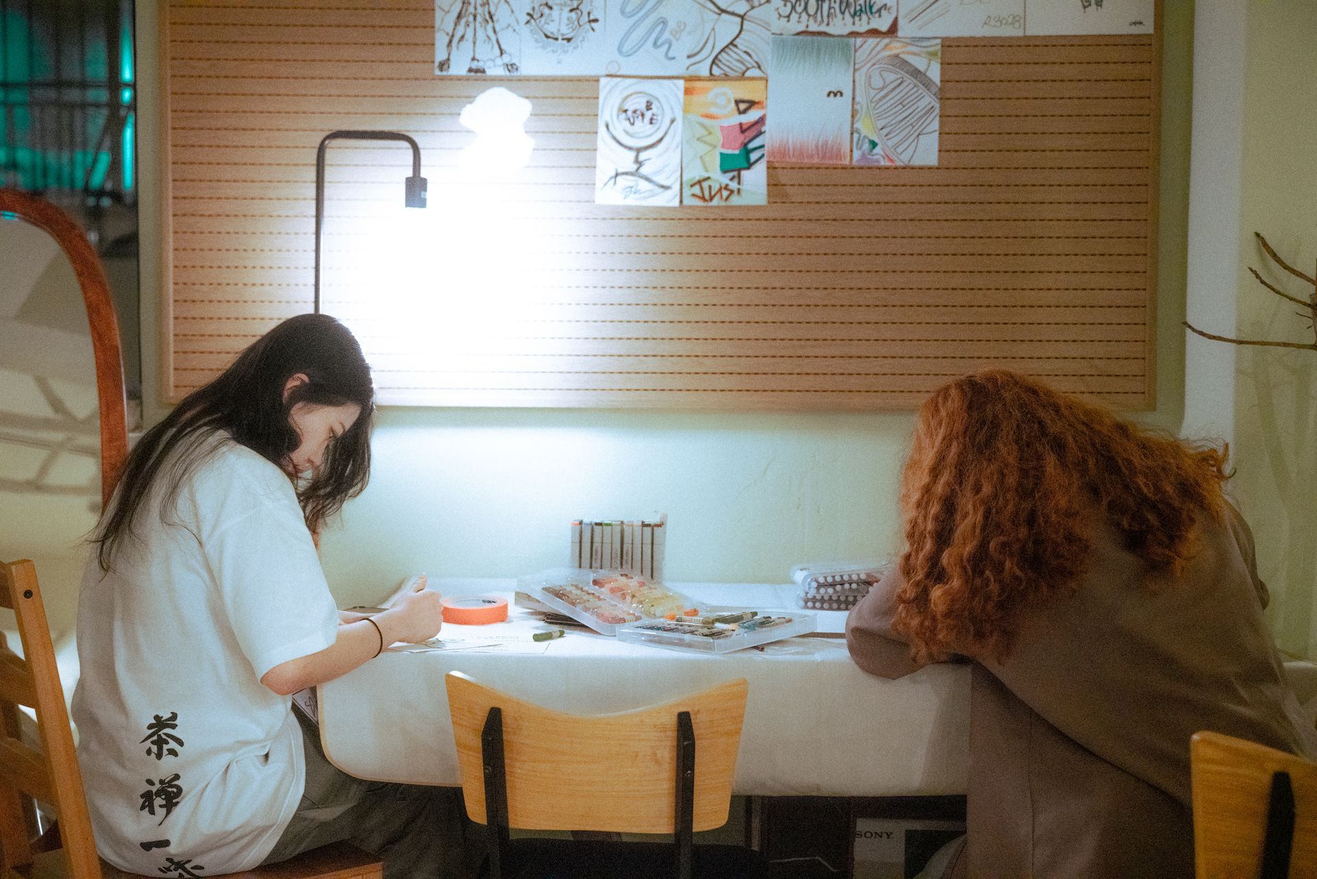 Two women seated at a table, drawing under a bright lamp.  A bulletin board of artwork hangs behind them.