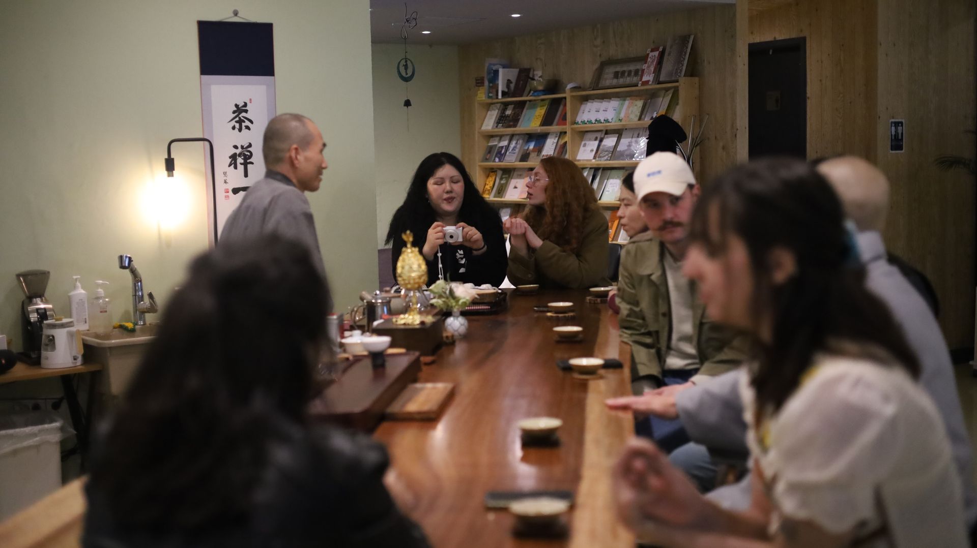 People sit around a long wooden table, drinking tea. A man in grey speaks. Tea ceremony in a room with books.