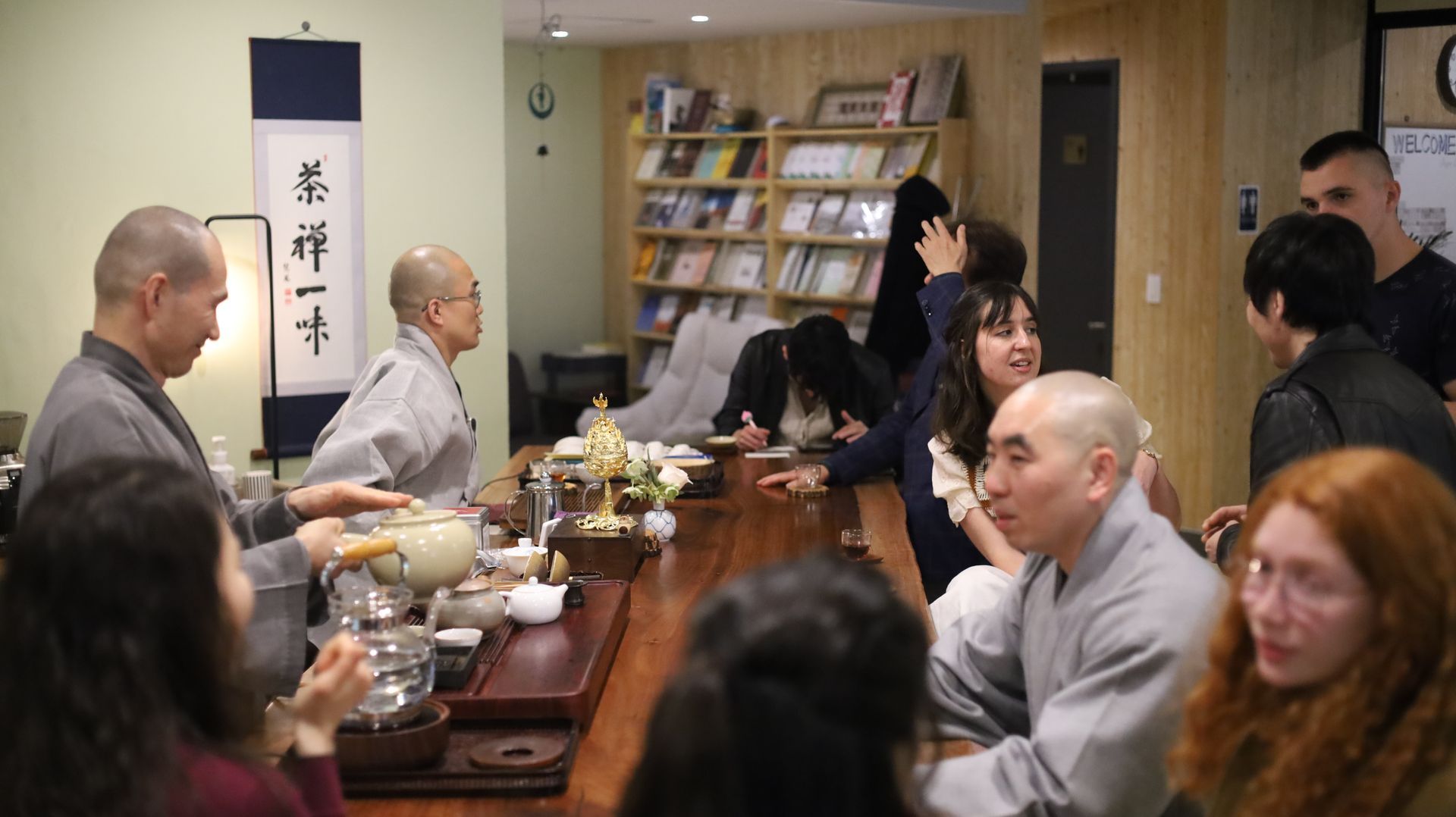 People seated at a long table, drinking tea. Two monks in gray robes serve. Interior with books, scroll.