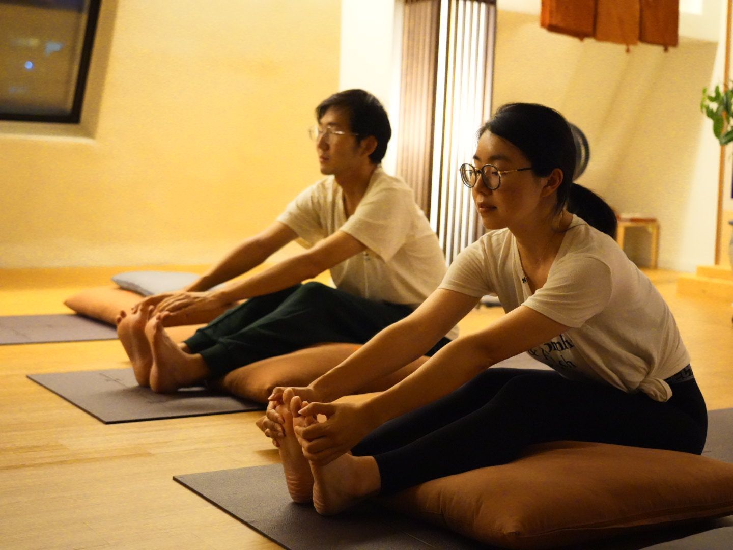 Two people stretching forward in yoga pose, indoors, on mats.