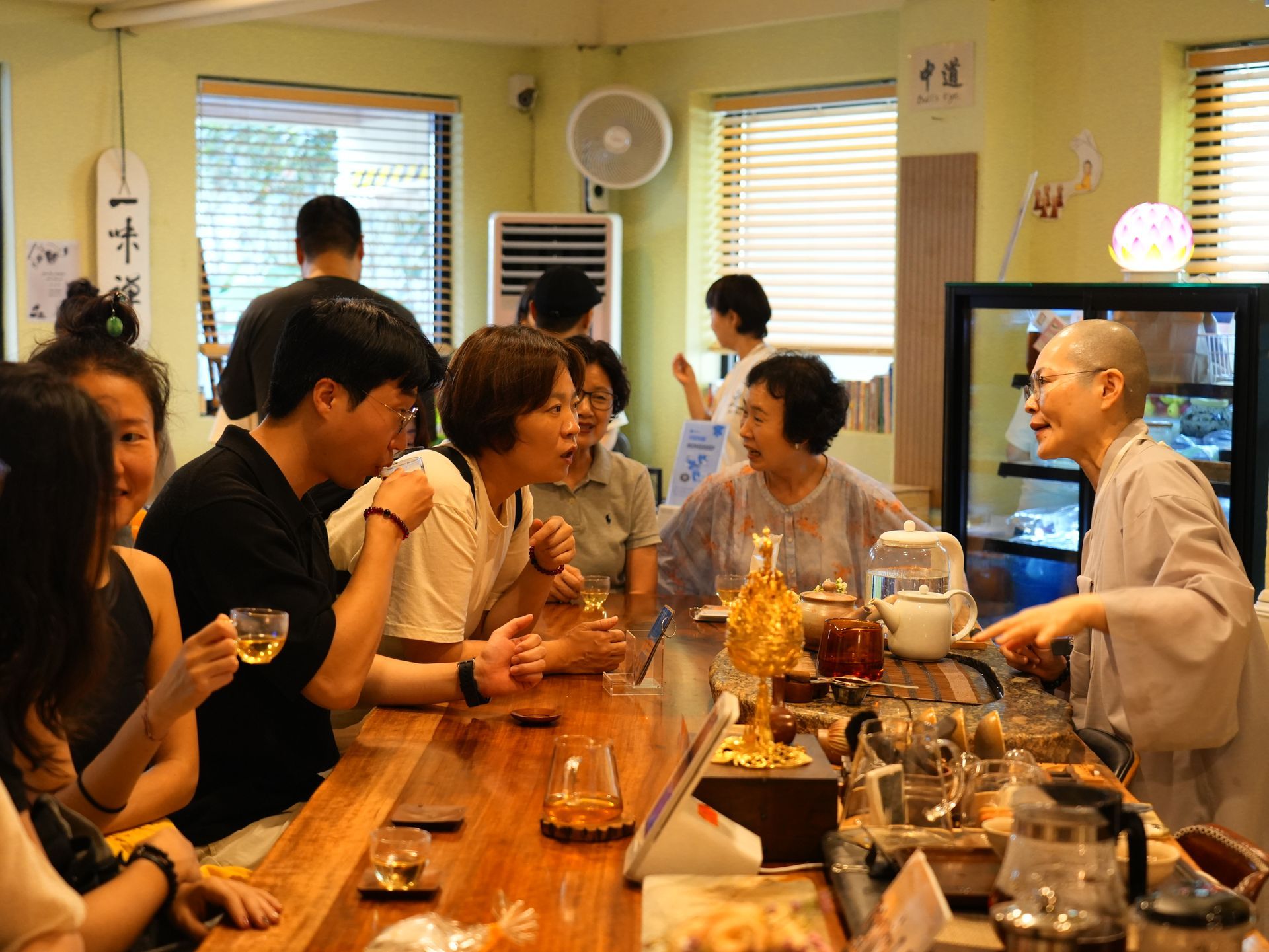 People chatting and drinking tea at a counter with a nun in a tea shop; bright interior with wooden surfaces.