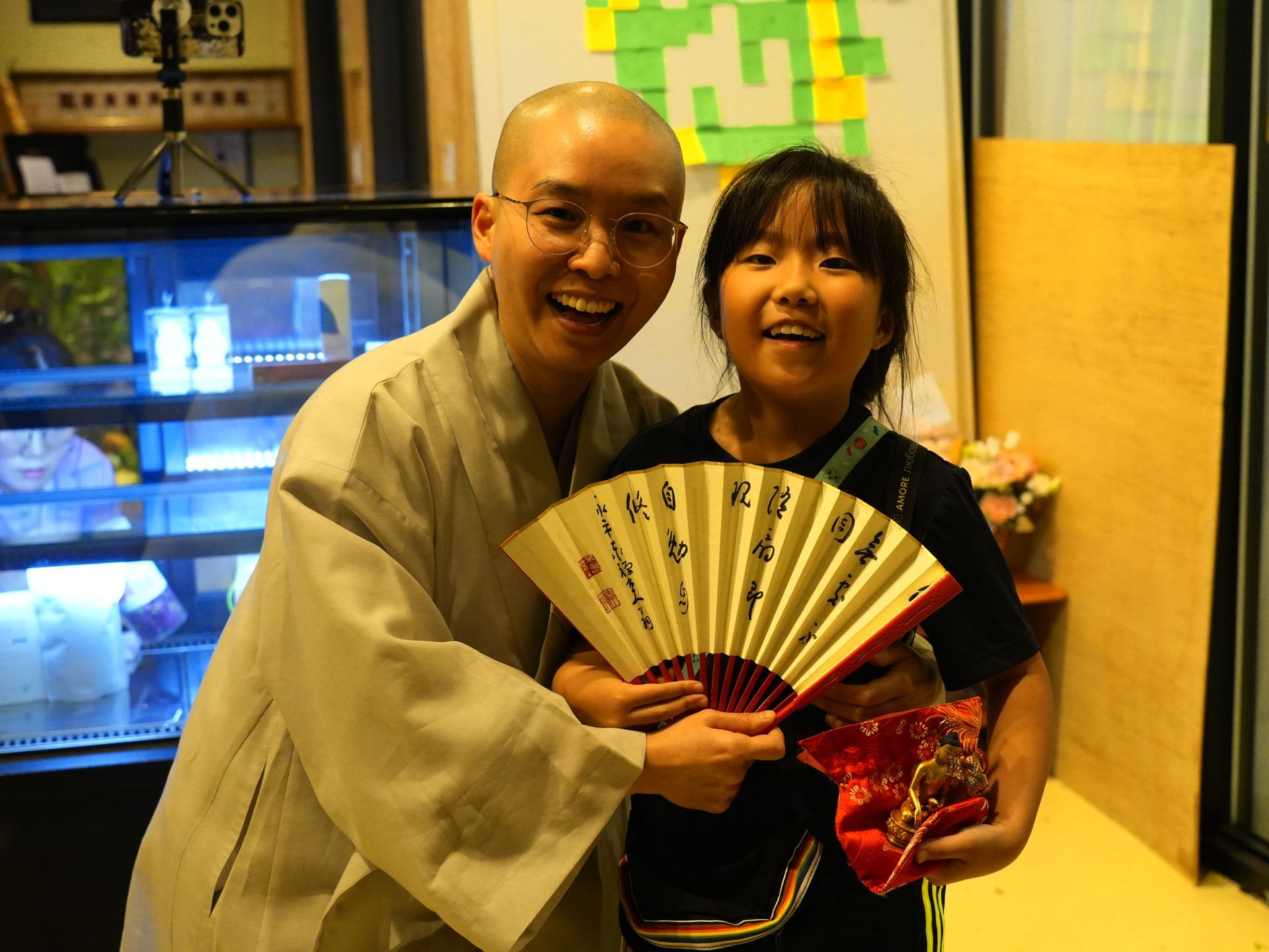 Man in robes and girl holding a fan, smiling together indoors. The man is bald.