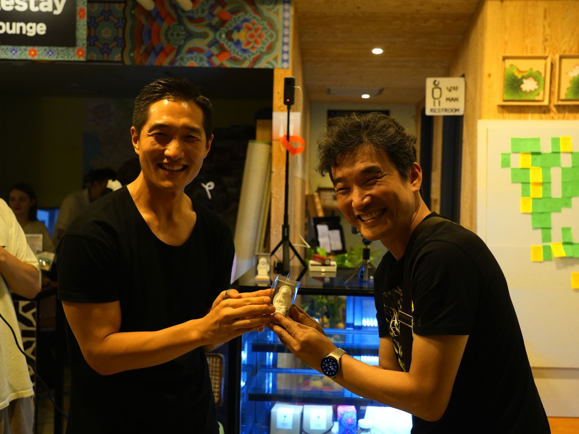 Two men smiling, one passing a small object. Interior setting, bright lighting.