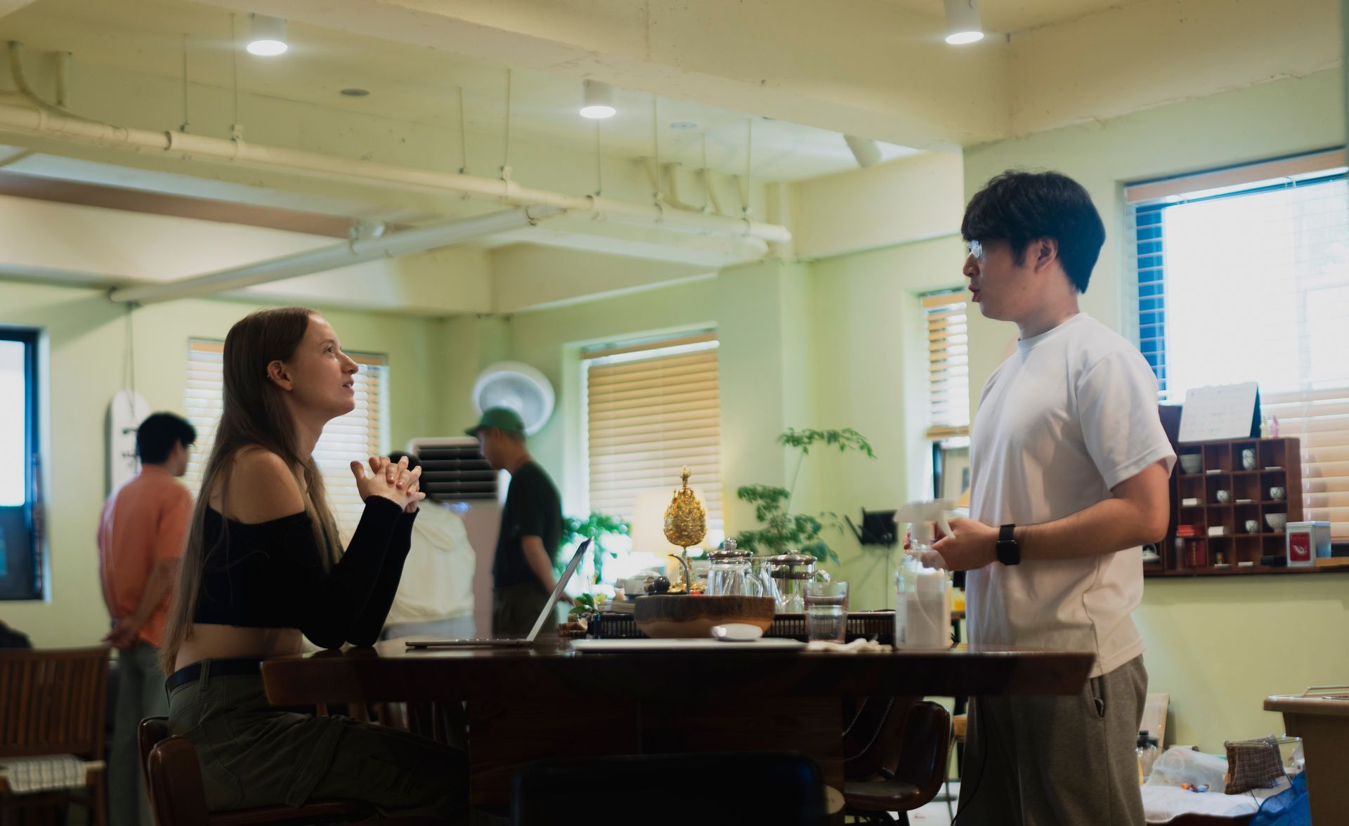 A woman gestures while talking to a man at a table in a cafe. Others in the background.