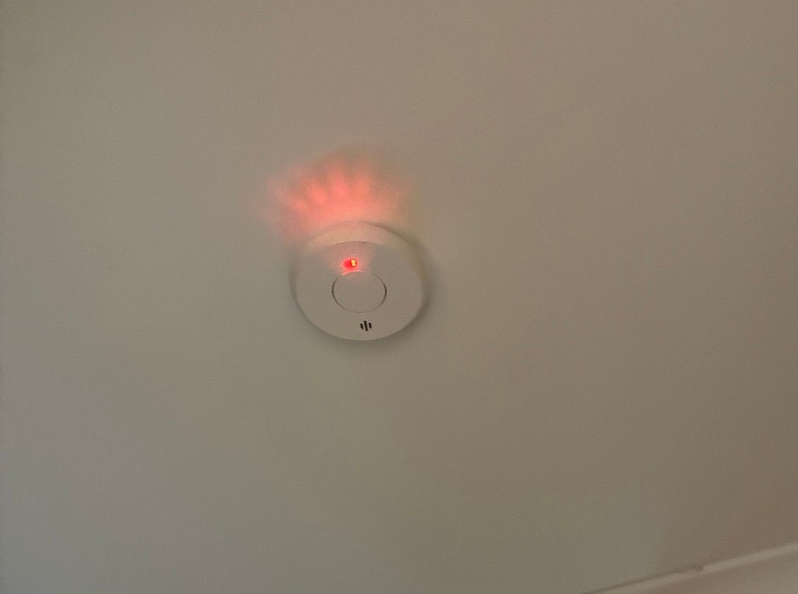 White smoke rises from a ceiling-mounted smoke detector against a gray backdrop.