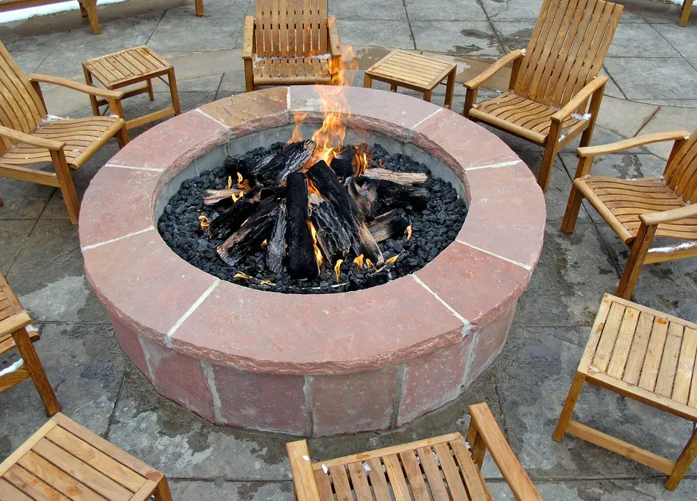 A fire pit with burning logs surrounded by wooden chairs on a patio.