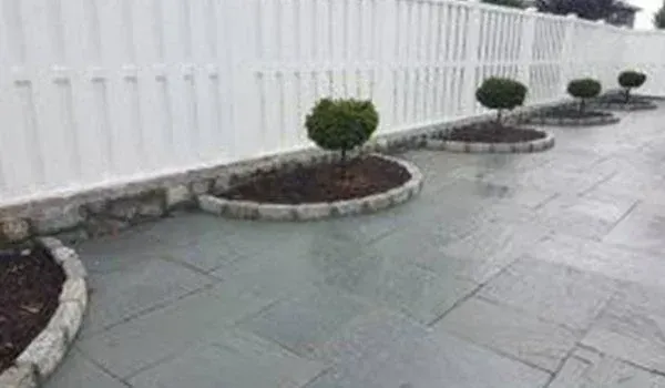 White fence bordering a stone patio with small round trees planted in circular beds.