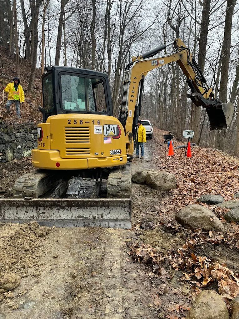A yellow Caterpillar excavator on a muddy path with workers. Forest in the background.