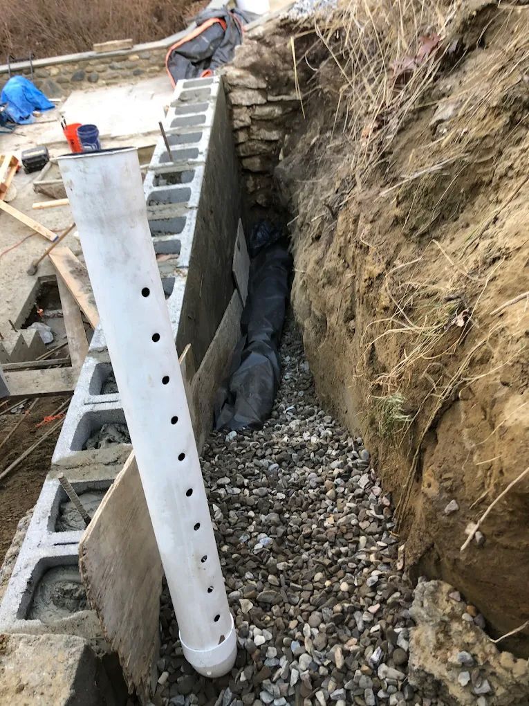 Construction of a retaining wall, with a drainage pipe in a gravel-filled trench. Workers are in the background.