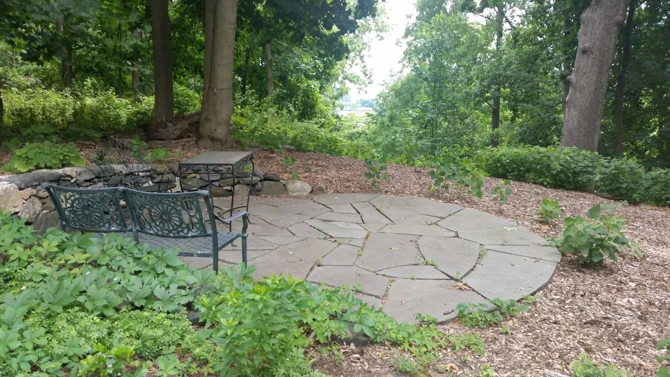 Stone patio with two benches, fire pit, and lush greenery in a wooded area.