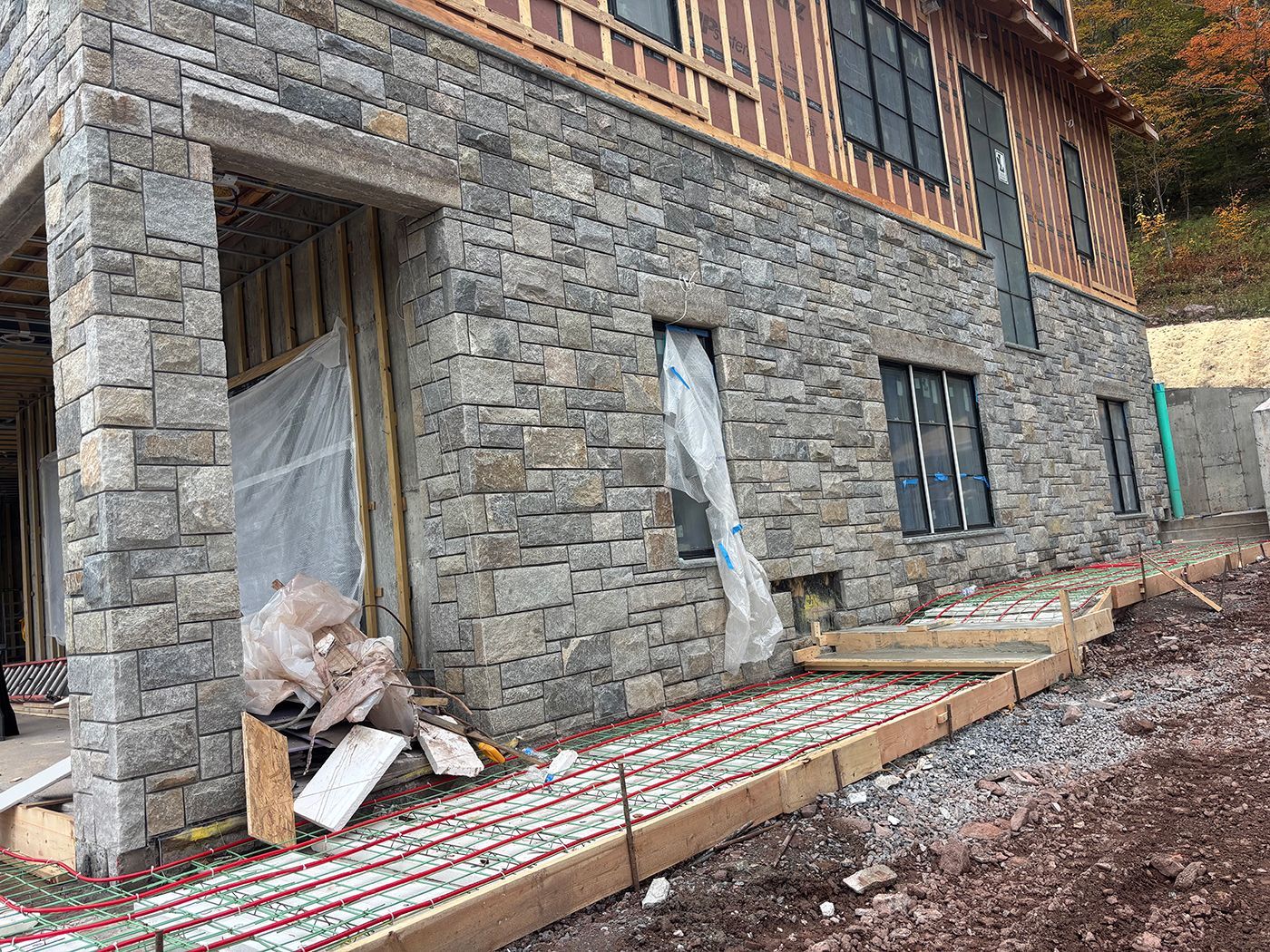 Stone-clad house under construction with exposed wood framing, windows, and a concrete pathway.