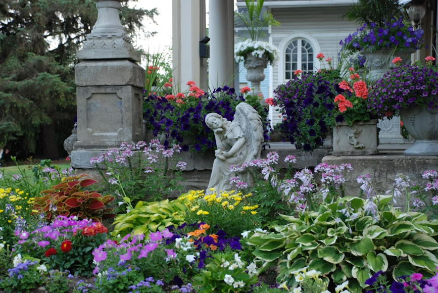 Stone angel statue amidst colorful flower garden and ornate porch.