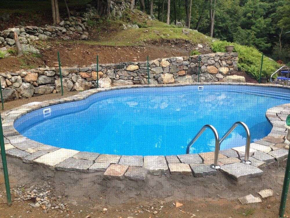 Swimming pool with blue water, surrounded by stone pavers and a retaining wall, set in a hillside.