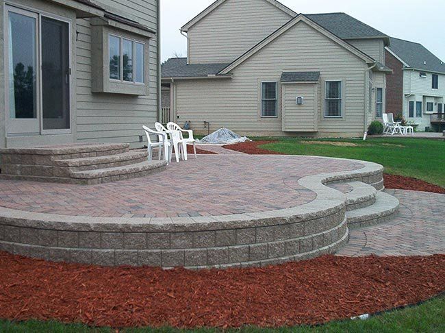 Brick patio with curved retaining wall, steps, and lawn; chairs sit near sliding glass door.