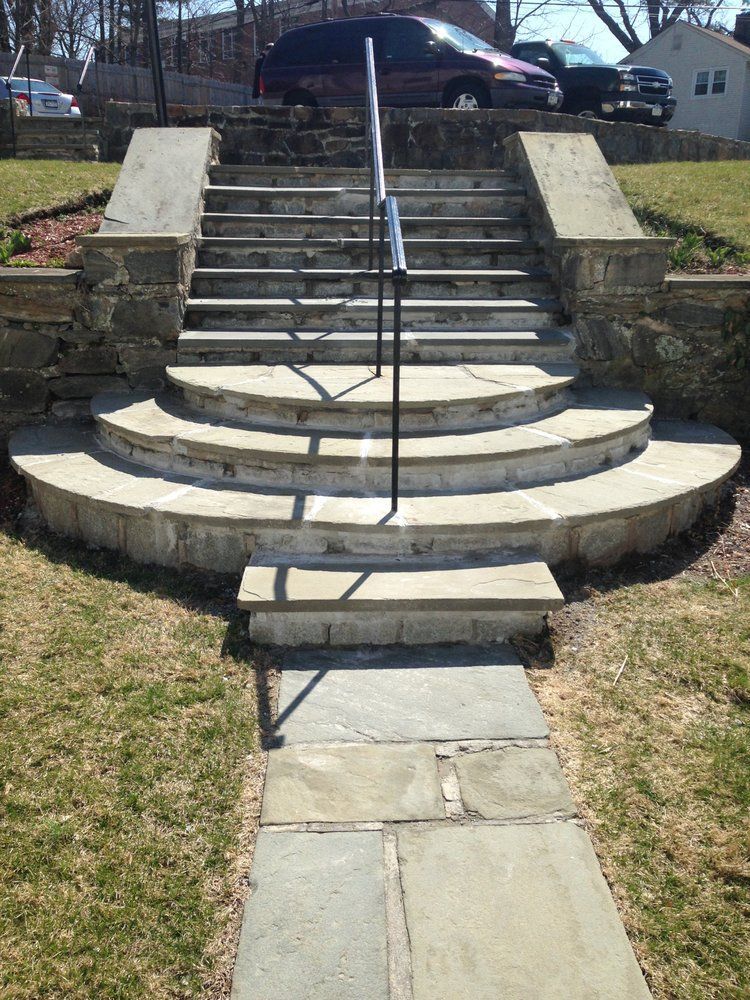 Stone steps leading uphill with a handrail. Pathway in front. Grassy yard with parked cars in the background.