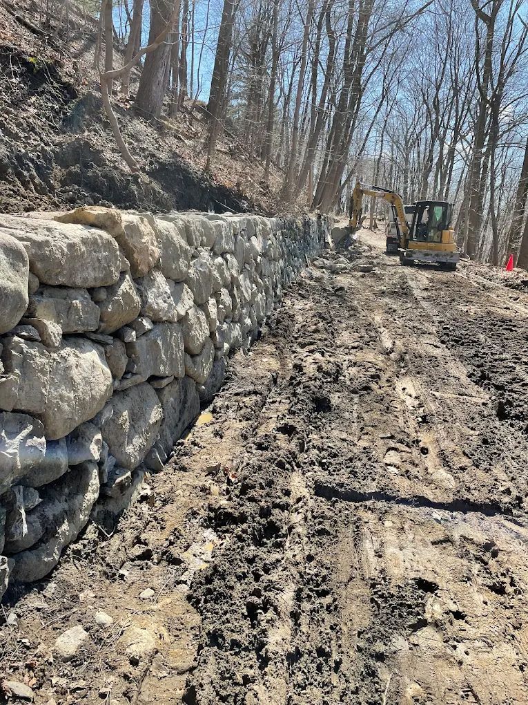 A stone retaining wall under construction along a muddy forest road. An excavator sits in the background.