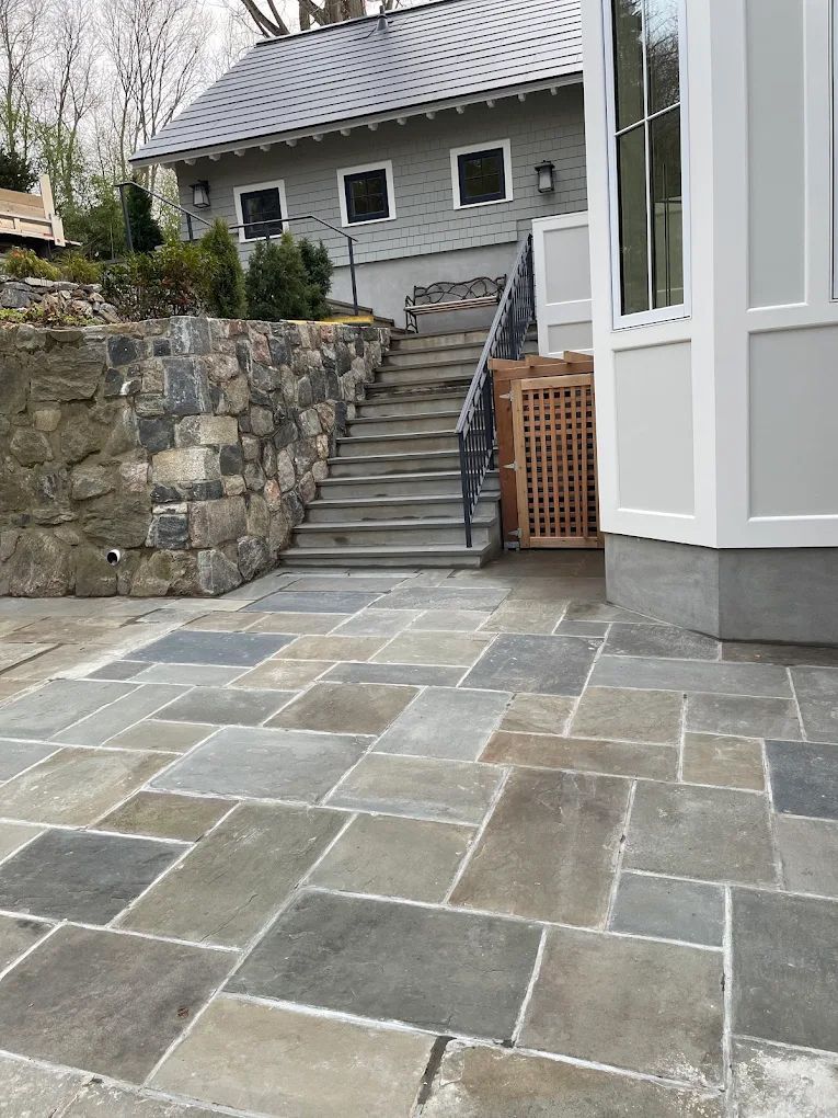 Stone patio and stairs lead to a house with gray siding. A stone wall is to the left.
