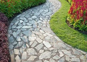 A curved stone pathway winds through a garden, bordered by vibrant red plants and green lawn.
