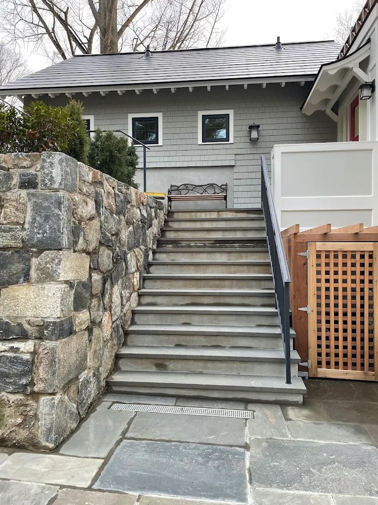 Stone stairs leading up to a gray building with a dark roof, alongside a stone wall and a wooden fence.