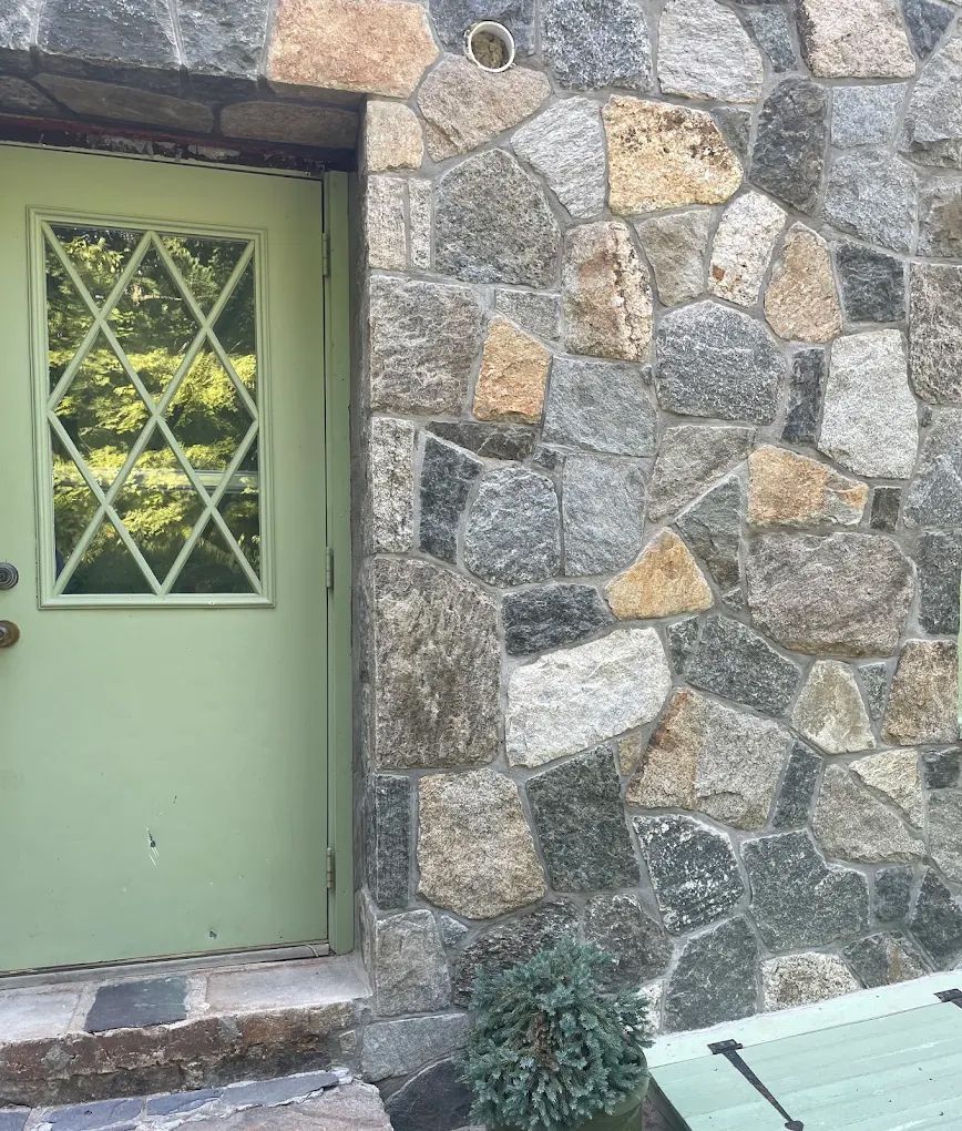 Green door with diamond-patterned window beside a multi-colored stone wall. A small bush sits at the base.