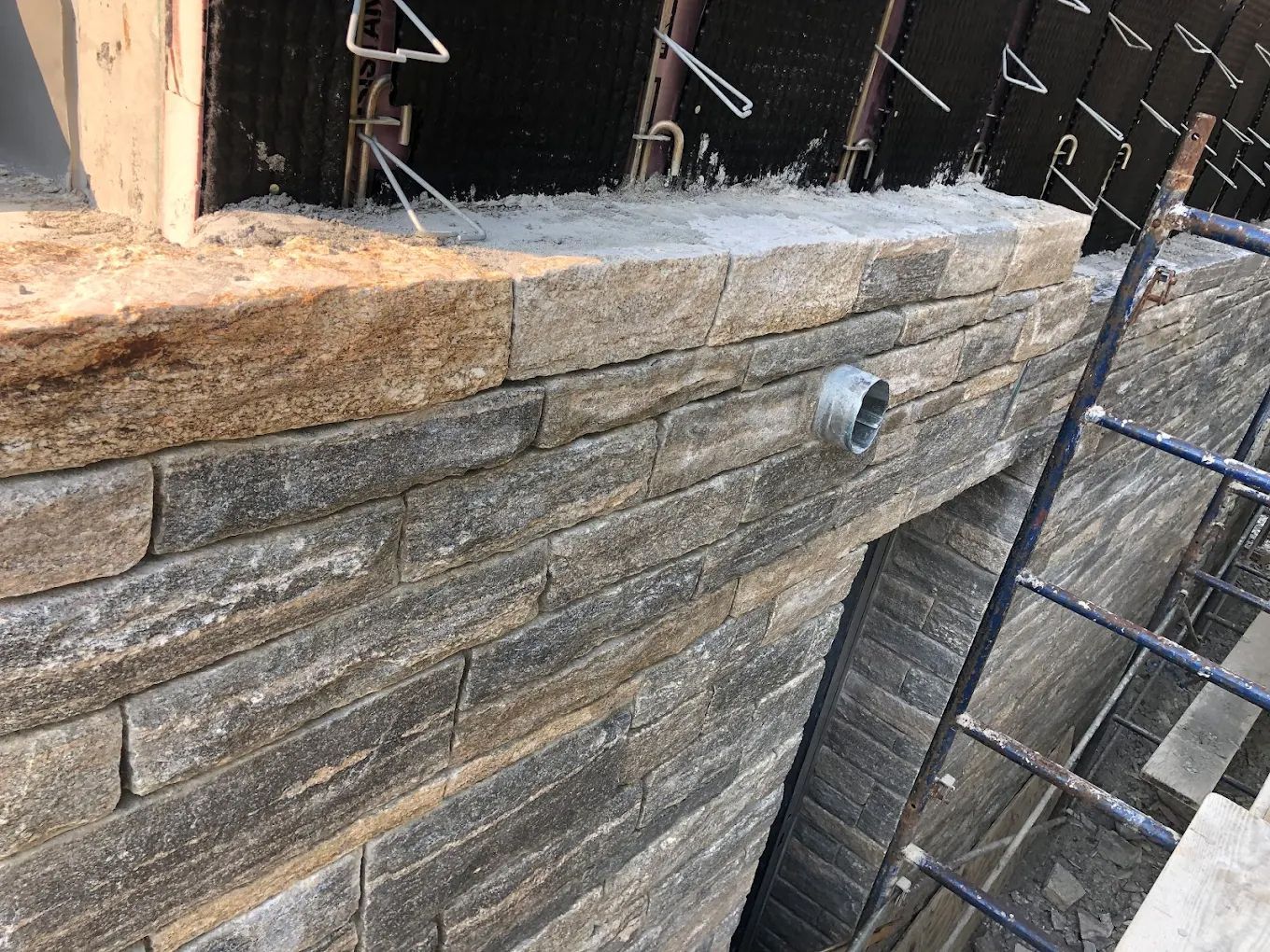 Stone brick wall under construction; grey and tan bricks, metal support, and pipe visible.