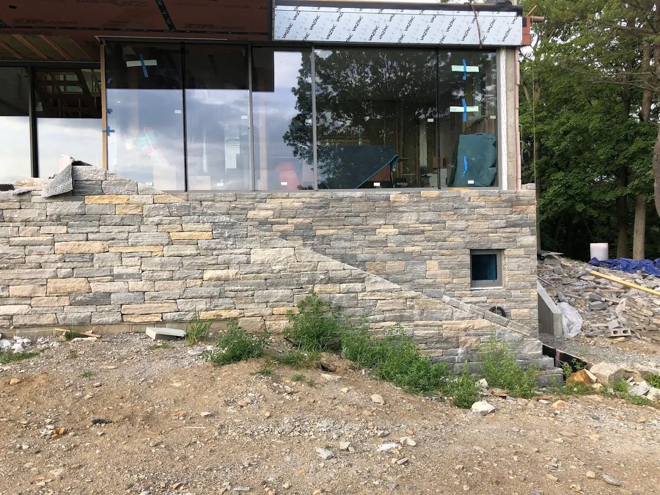 Stone-clad building under construction with large windows and a small square window, set on a gravelly slope.