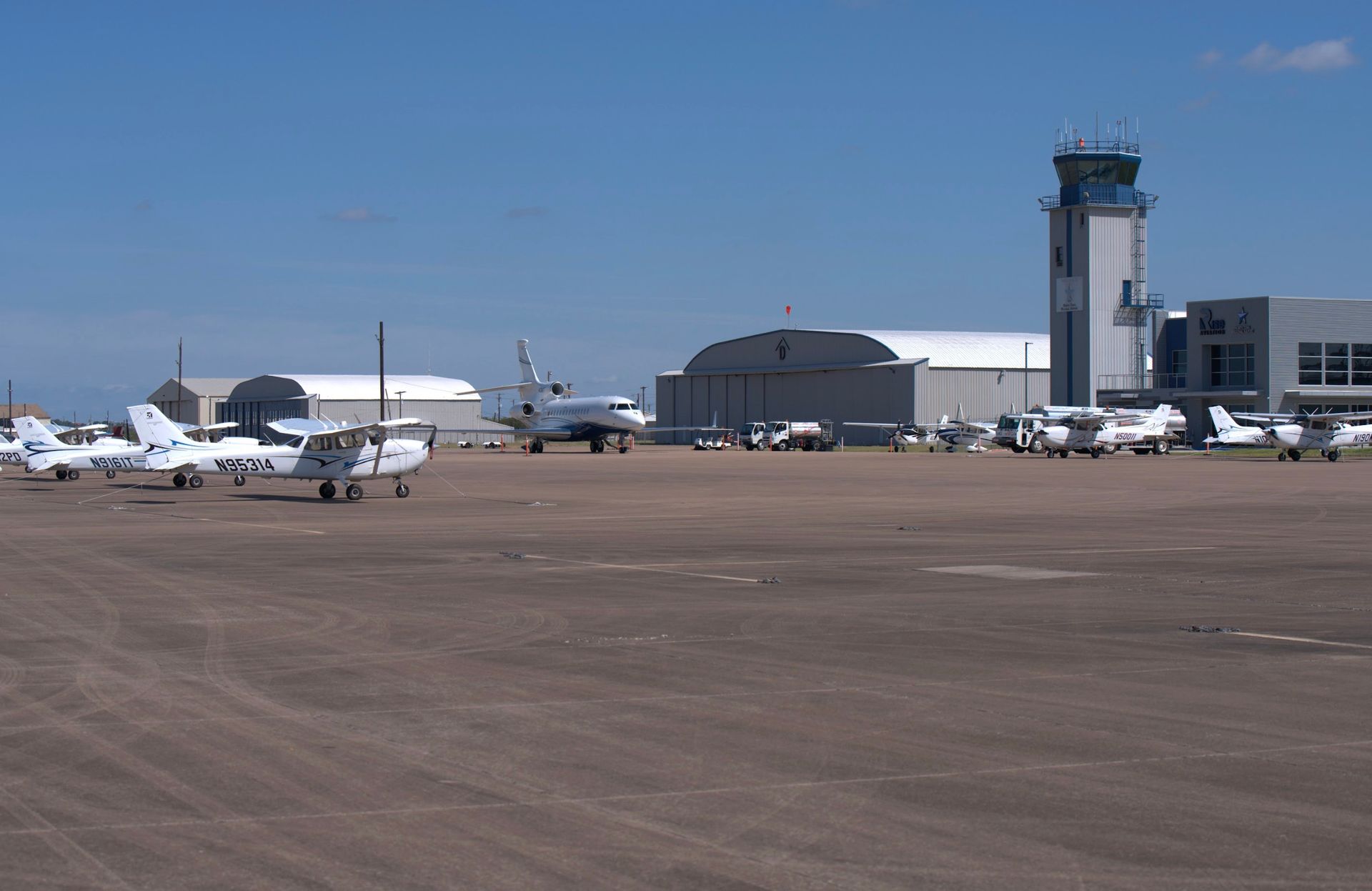 Airfield scene: Planes on a tarmac, control tower at right, hangars in background under a blue sky.