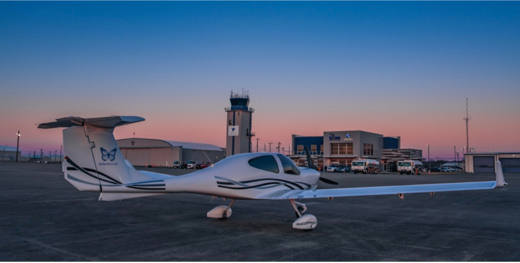 White airplane on a tarmac with a control tower and building in the background at sunset.