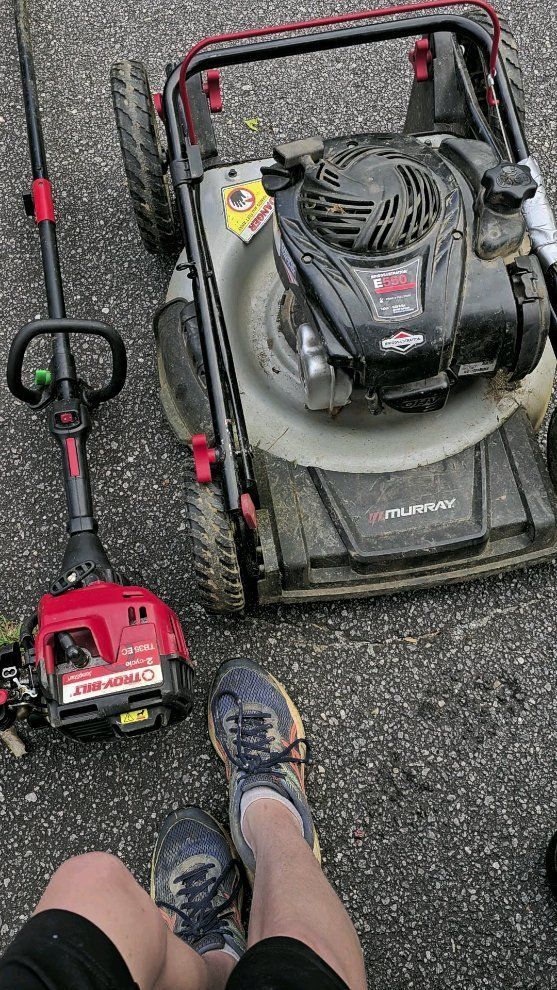 A person is sitting next to a lawn mower and a trimmer.