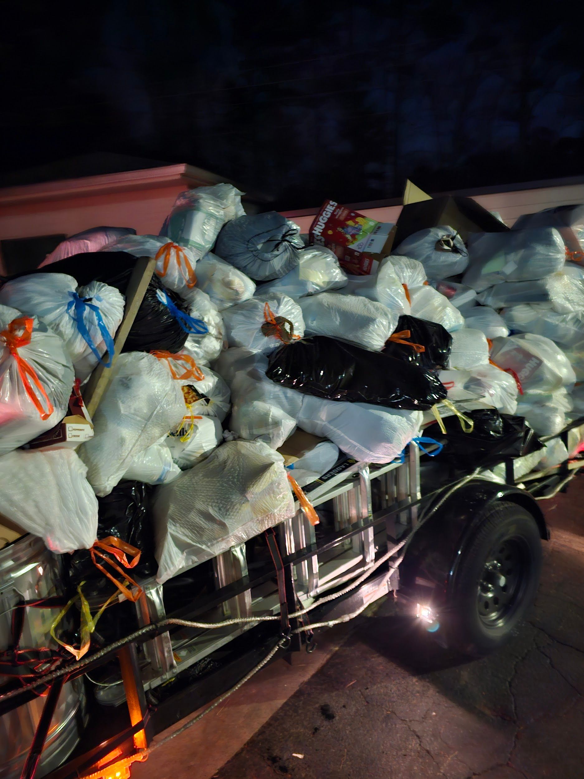 A trailer filled with bags of trash is parked on the side of the road at night.