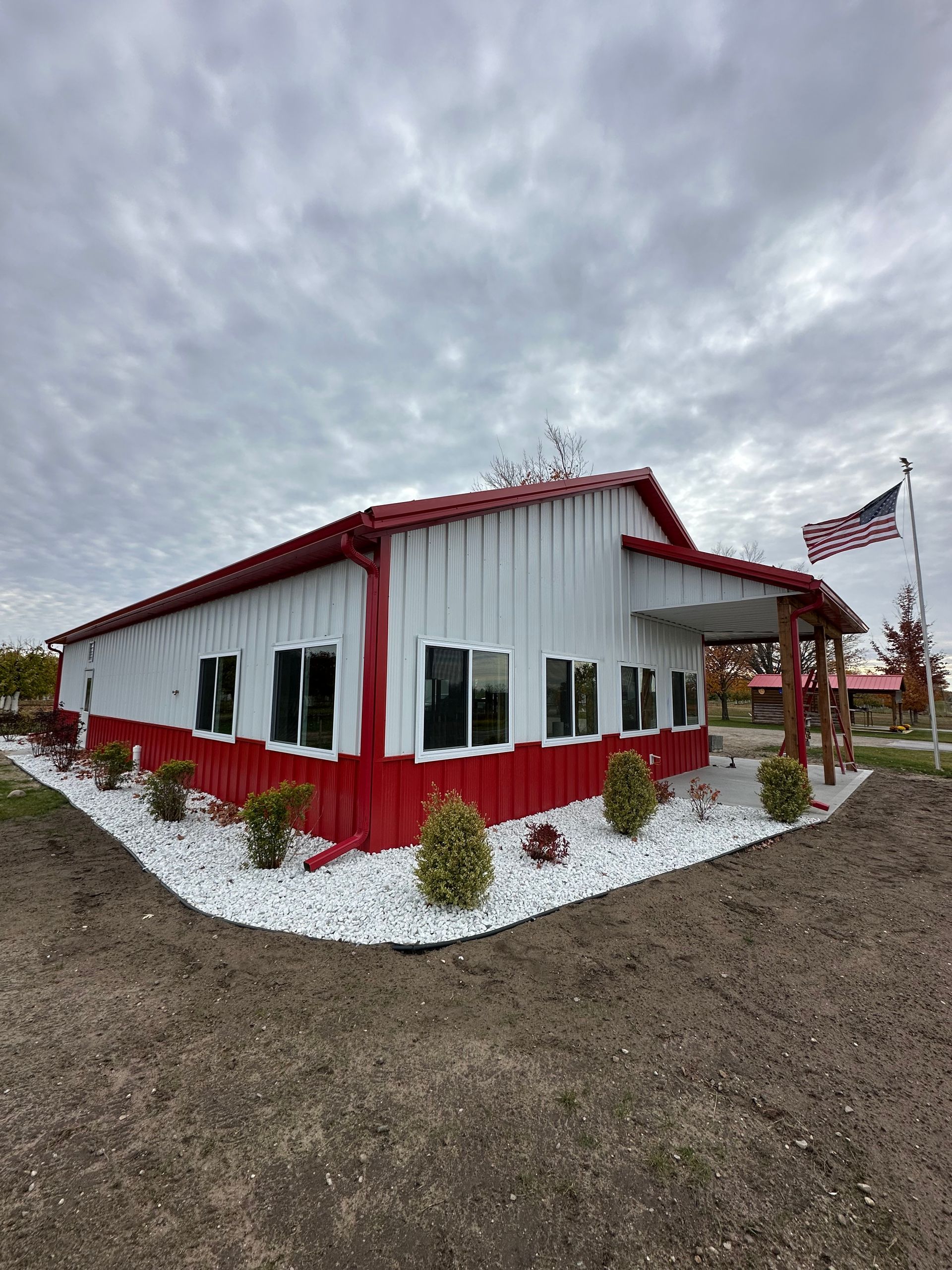 A white and red metal building with windows and a small front porch, surrounded by white landscaping rocks and shrubs.