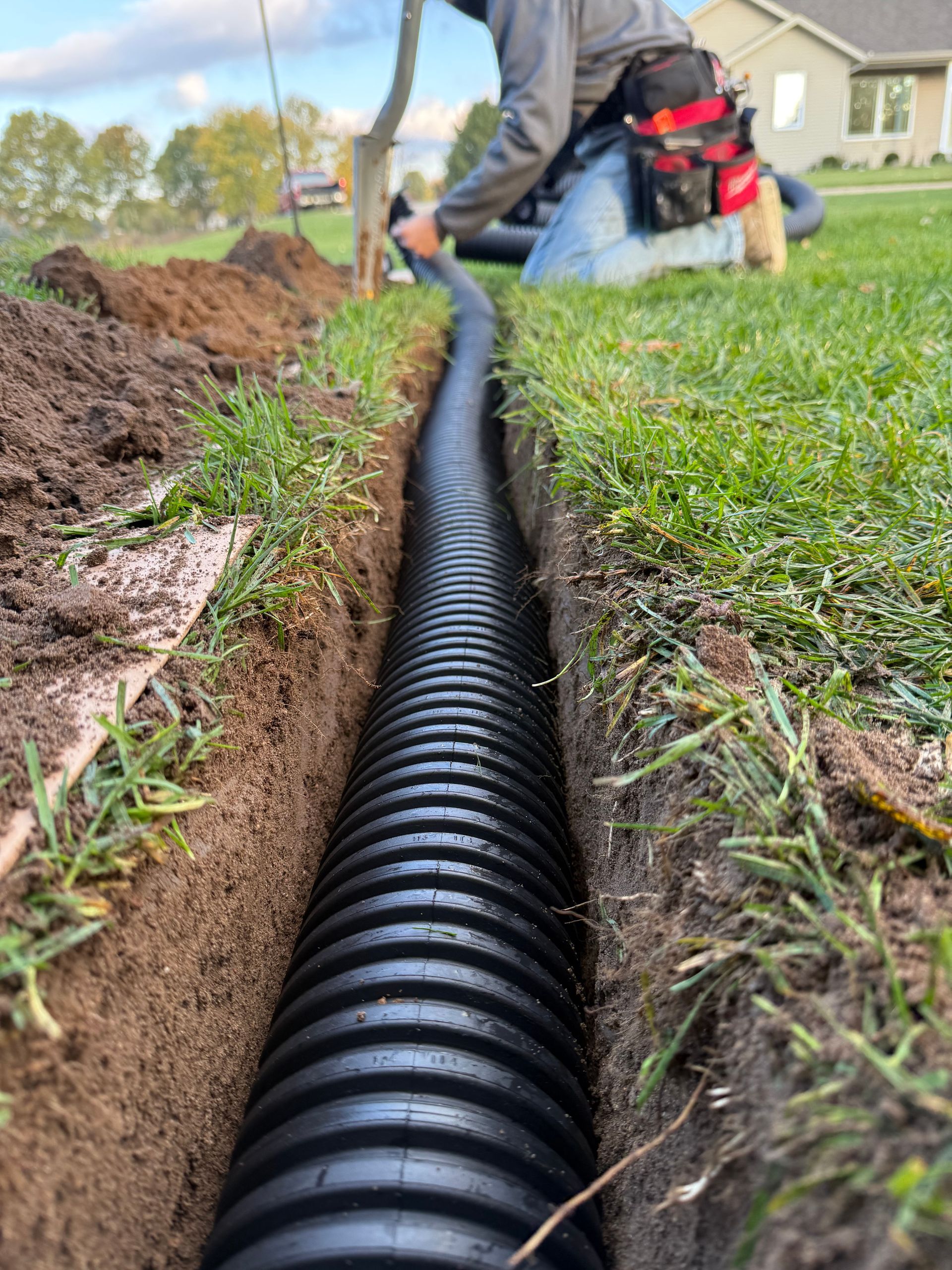 A person installing a black corrugated drainage pipe into a narrow trench in a grassy yard.