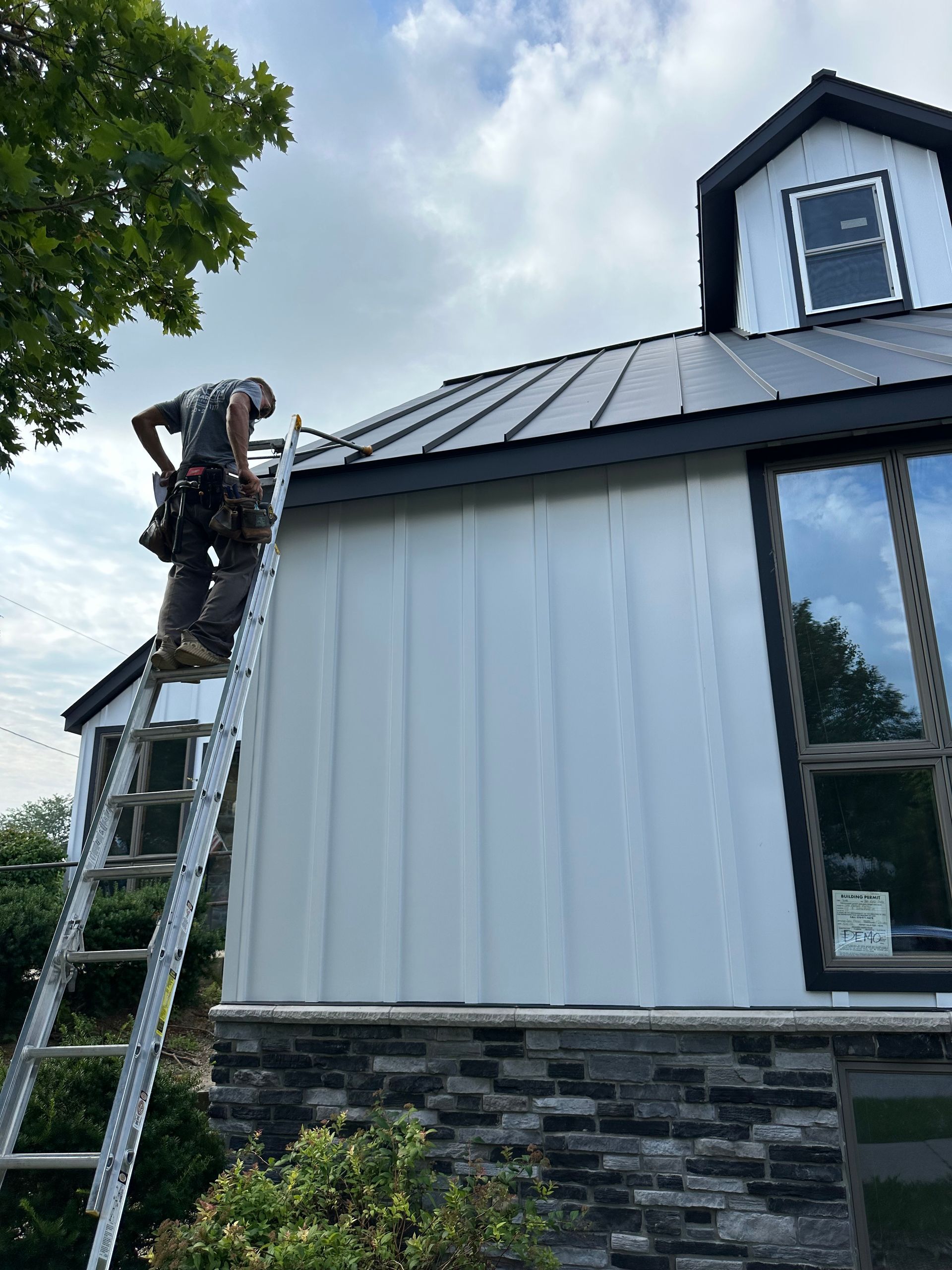A worker on a ladder installs flashing on the roof of a modern house with white vertical siding and stone foundation.
