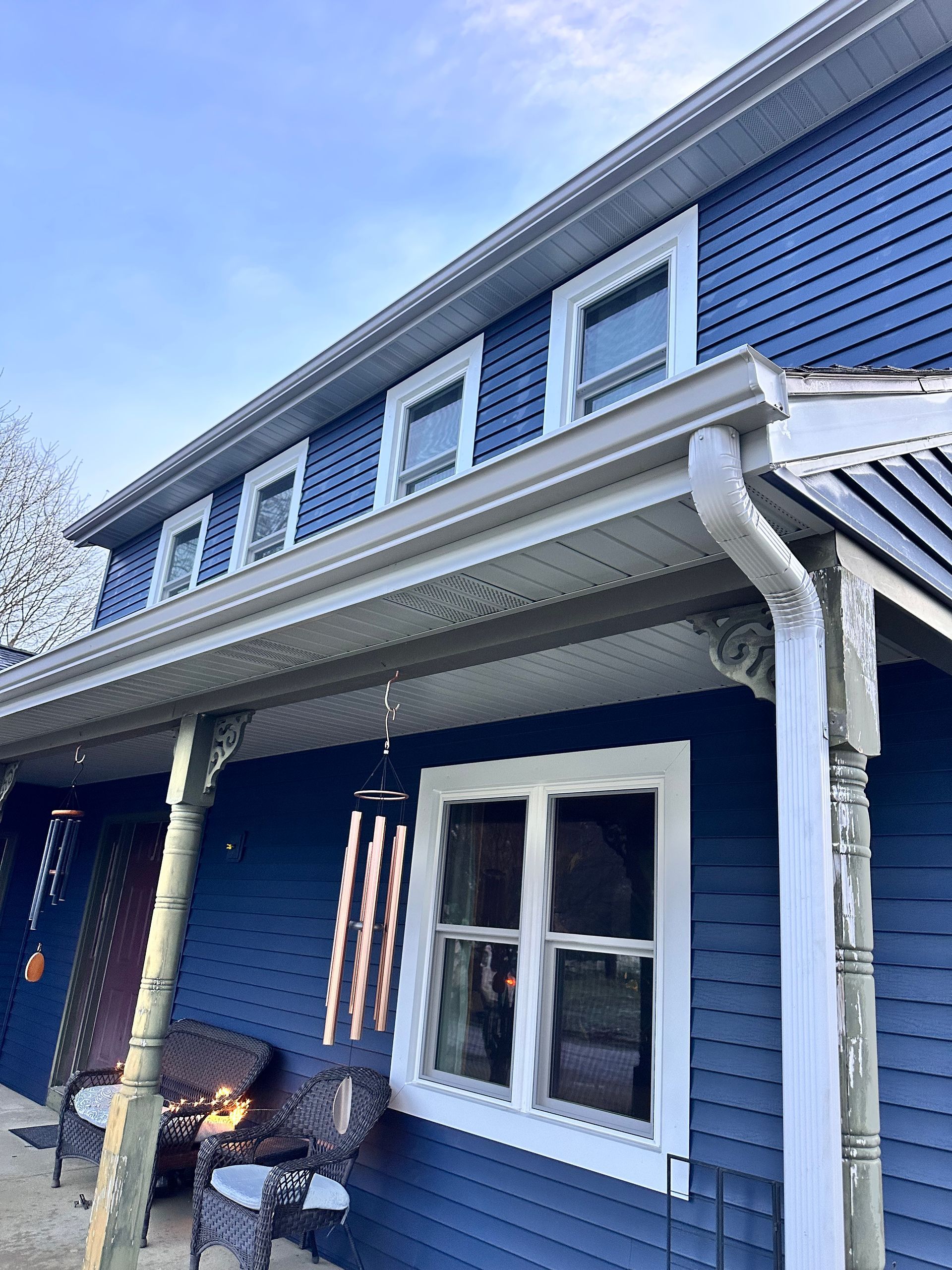A blue two-story house exterior with a covered porch, white trim, and hanging wind chimes under a blue sky.