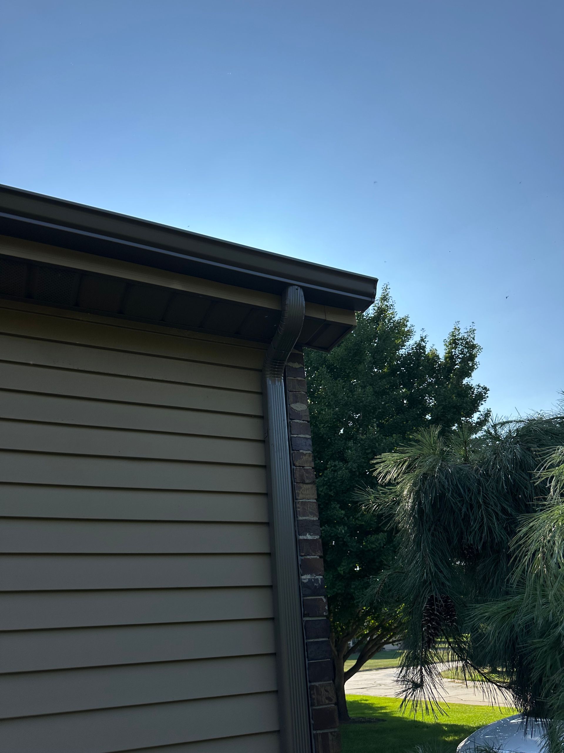 Exterior view of a house corner showing beige horizontal siding, a dark roof edge, a gutter downspout, and leafy trees.