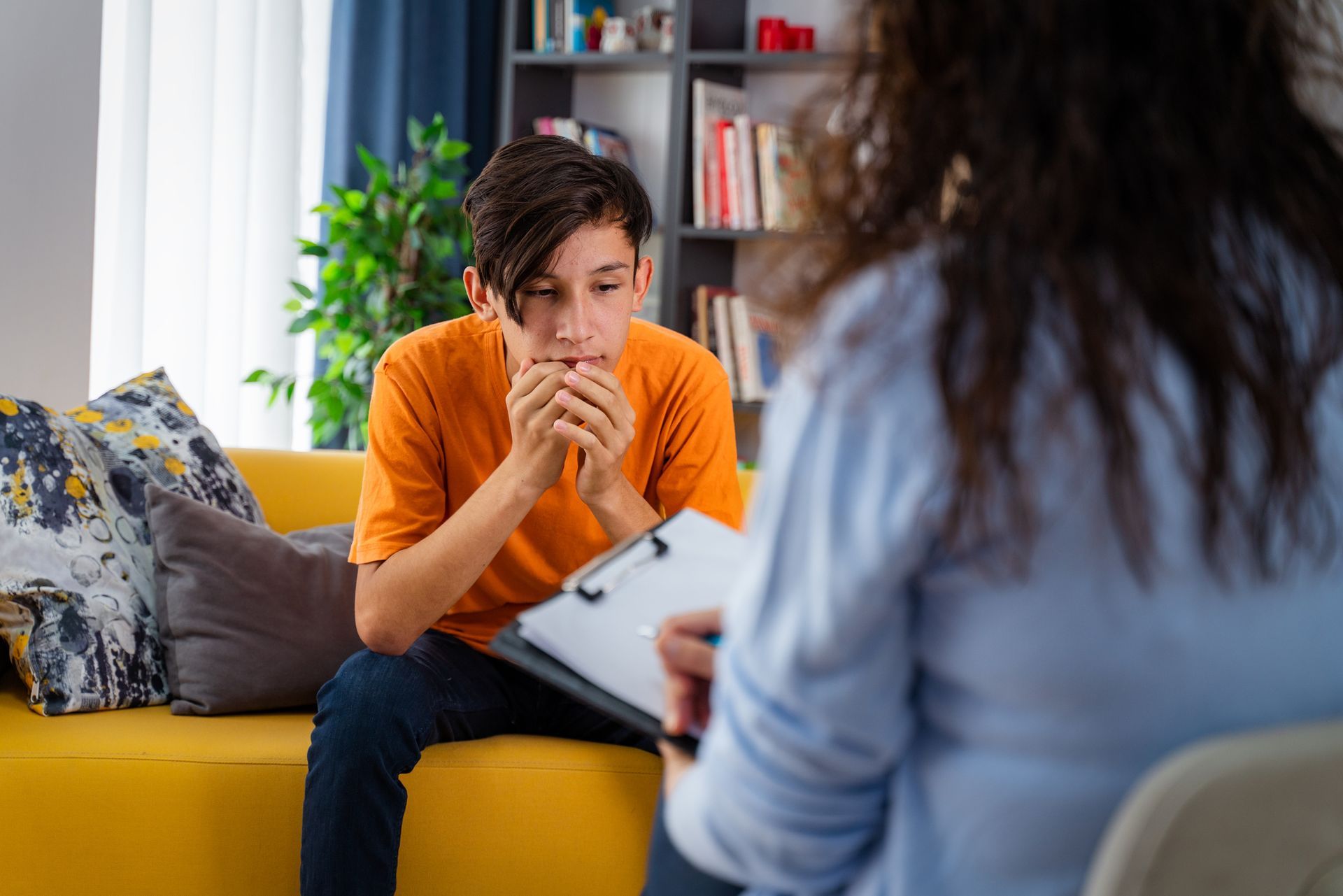 A young boy is sitting on a yellow couch talking to a woman.