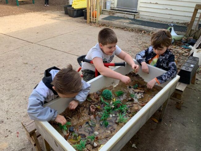 Three young boys are playing in a sandbox with toy dinosaurs.