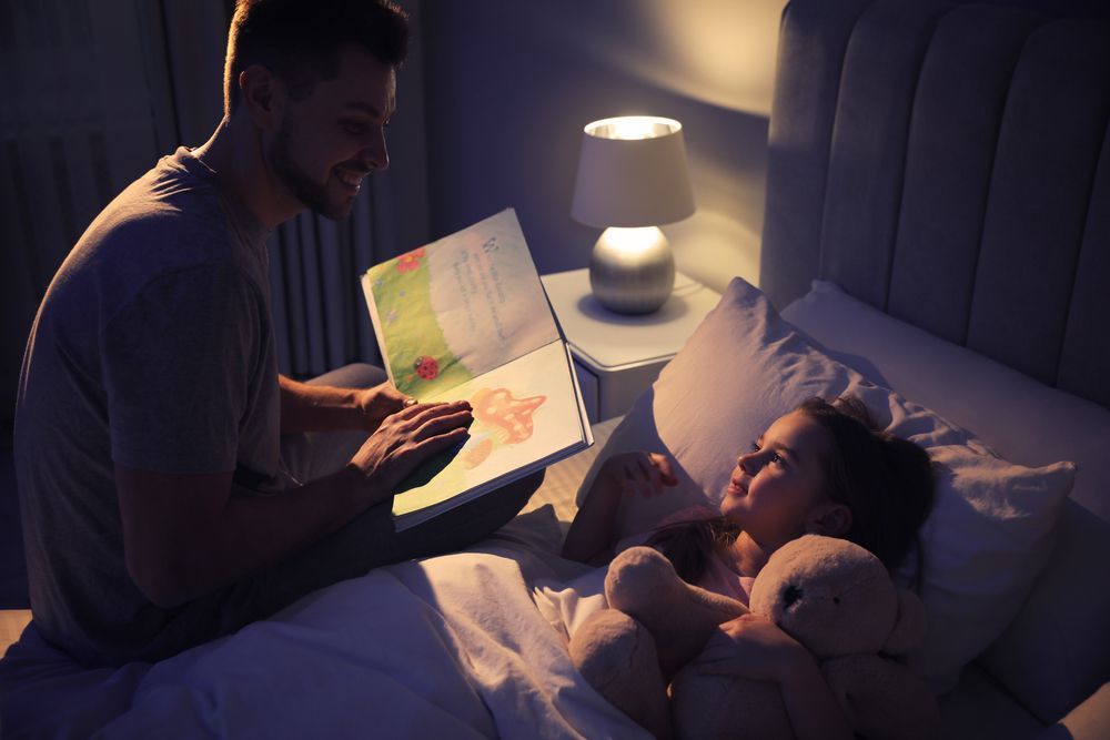 A man is reading a book to a little girl in bed.