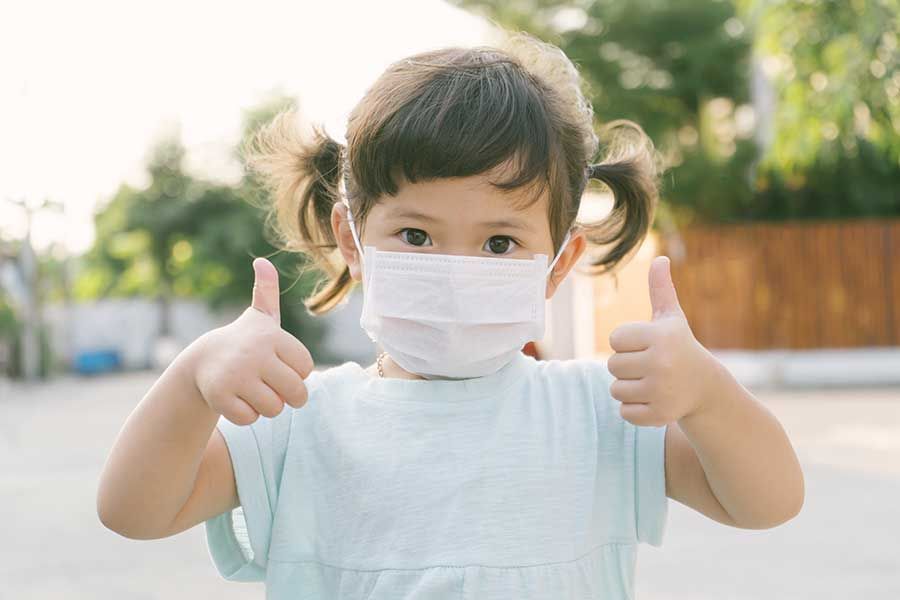 A little girl wearing a face mask is giving a thumbs up.