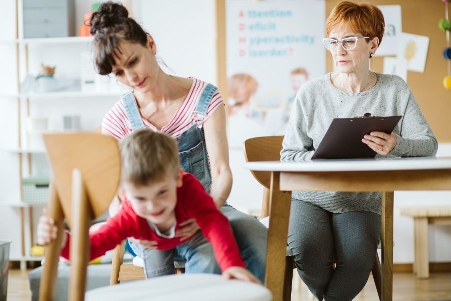 A woman is sitting at a table with a clipboard and a child is crawling on a chair.